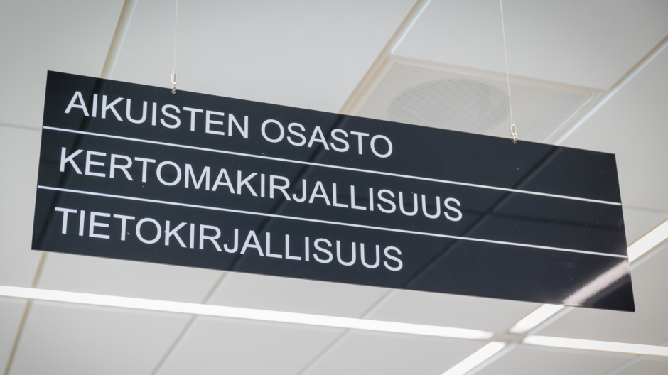 Kirjaston katosta roikkuva opaste kuvattuna alhaalta. Opasteessa lukee allekkain valkoisella mustalla taustalla aikuisten osasto, kertomakirjallisuus ja tietokirjallisuus.